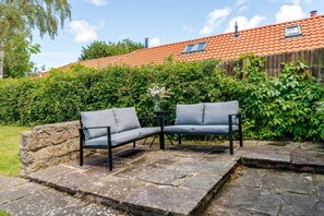 Une terrasse avec deux canapés gris et une petite table, entourée d’un dallage en pierre et d’un mur en pierre.
