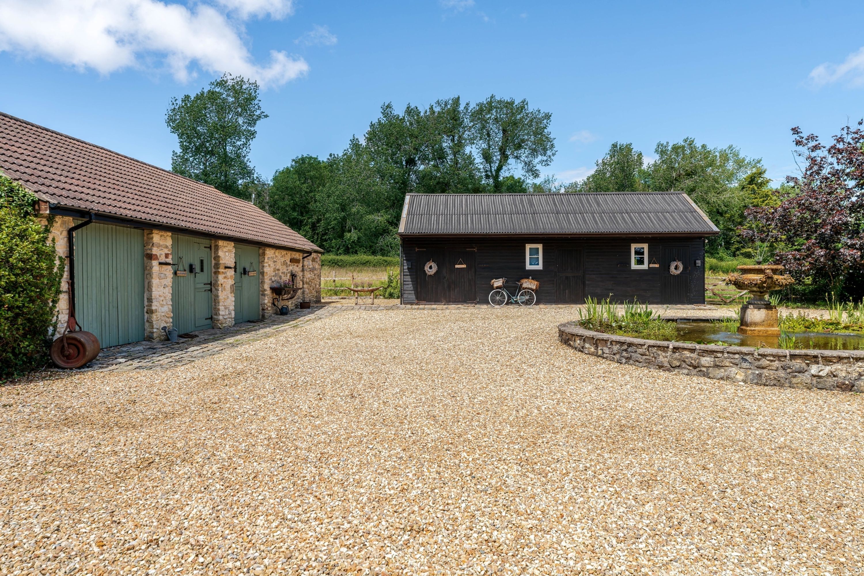 A gravel driveway leading to a black building with a metal roof, surrounded by greenery and a small pond.