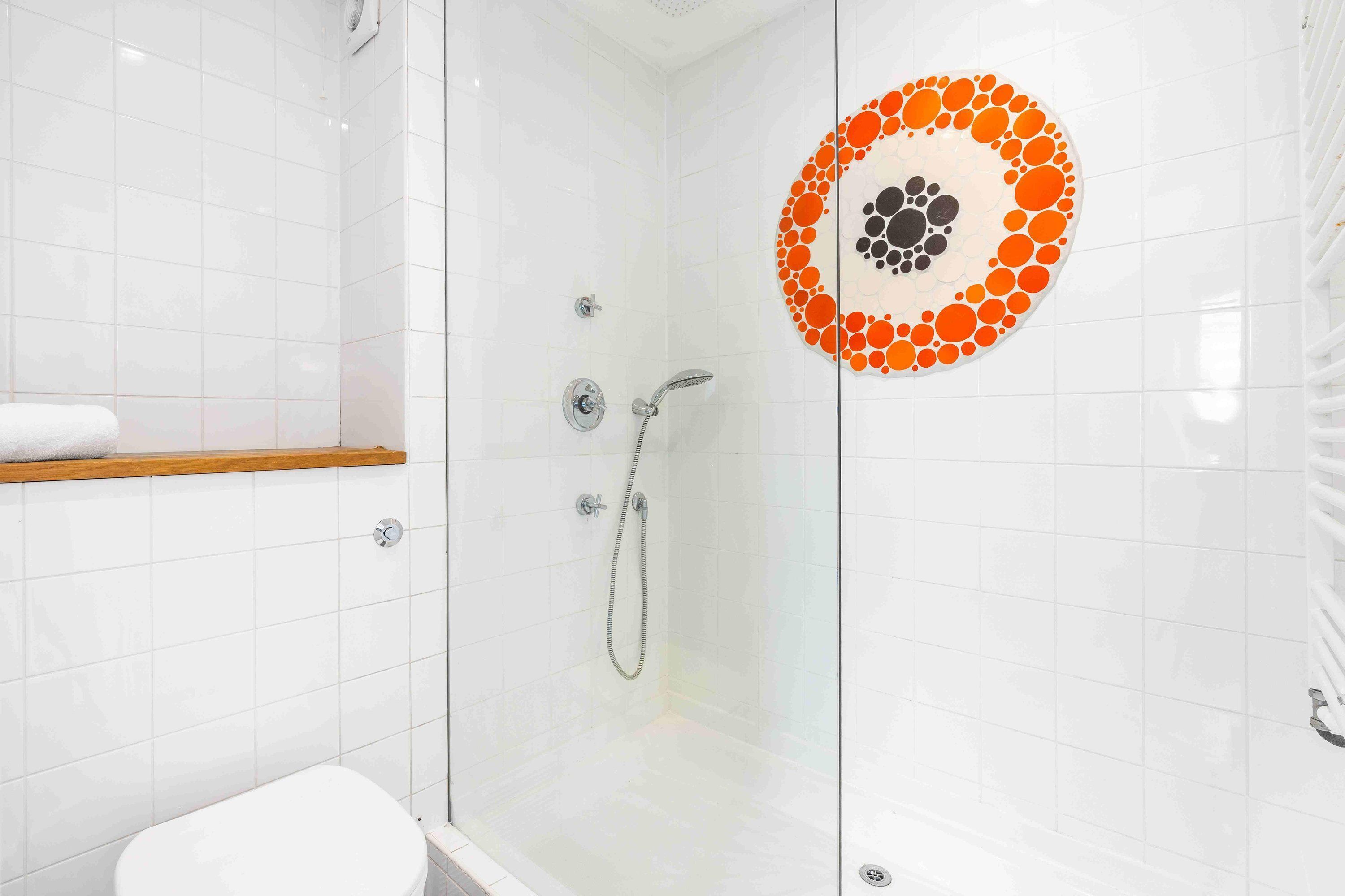 Una ducha con paredes de vidrio, pared de azulejos blancos, estante de madera y adorno circular de azulejos.