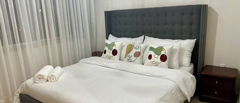 Un lit bien fait, avec du linge de lit blanc, une tête de lit rembourrée grise et des coussins décoratifs aux motifs abstraits.