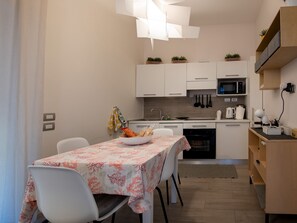 A compact kitchen with white cabinets, a microwave, and a sink. There is a dining table with chairs and a bowl of fruit. - Le Terrazze sul Cortile - Red (Salerno)