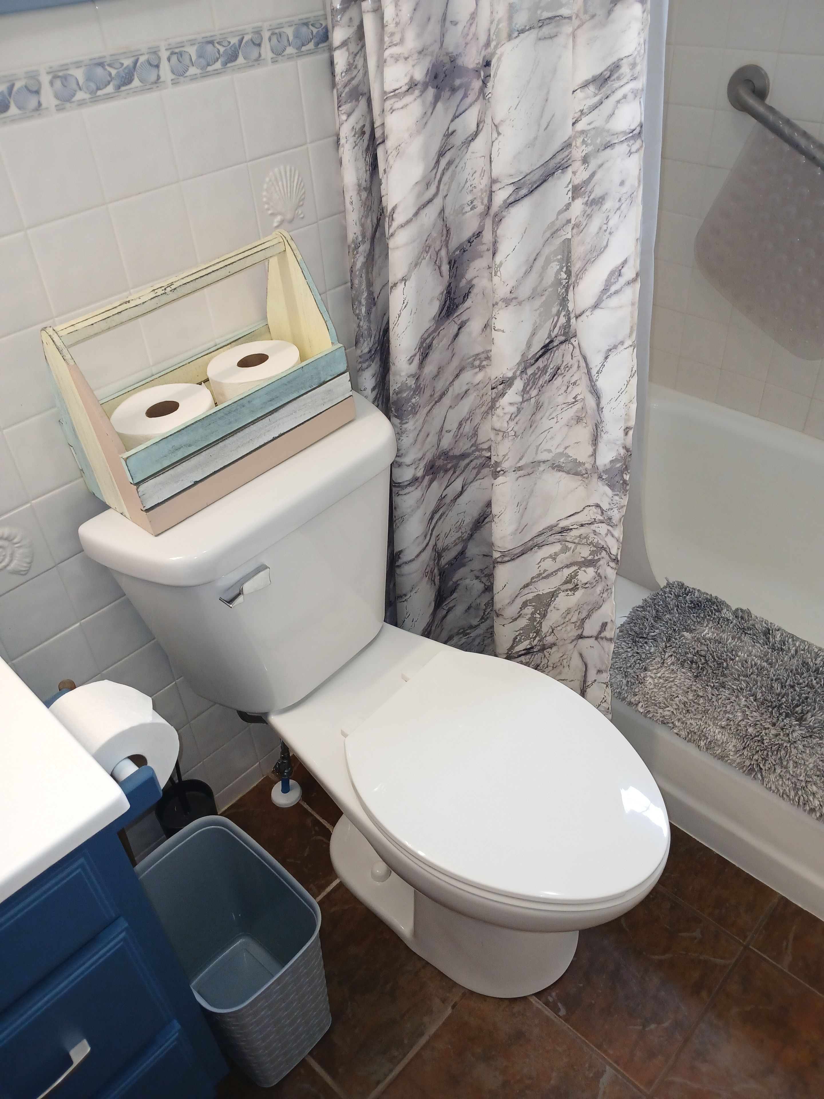 A bathroom with a toilet, a shower with a gray bath mat, and a blue trash bin.