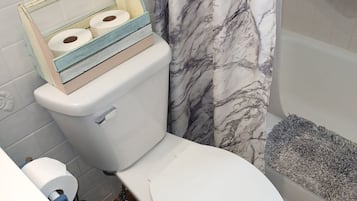 A bathroom with a toilet, a shower with a gray bath mat, and a blue trash bin.