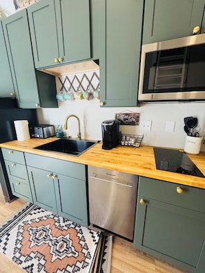 A compact kitchen with teal cabinets, a wooden countertop, a stainless steel dishwasher, and a coffee station.
