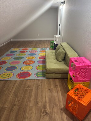 Et værelse med trægulv, en farverig legemåtte med bogstaver, en grøn sofa med puder og stablende plastikbokse i pink og orange.