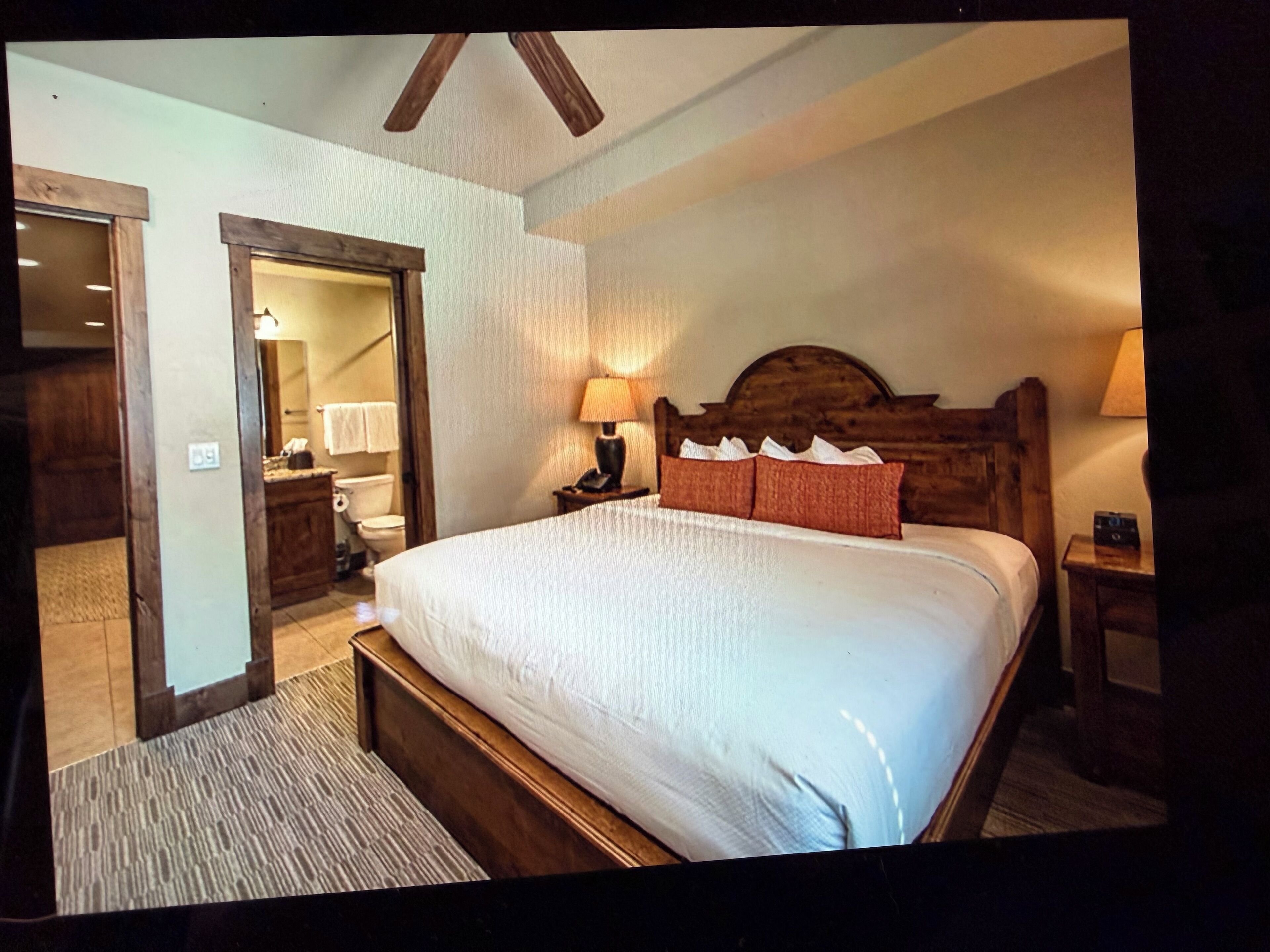 Ein Schlafzimmer mit einem hölzernen Bett, einem Nachttisch, einer Lampe und einem durch eine geöffnete Tür einsehbaren Badezimmer.