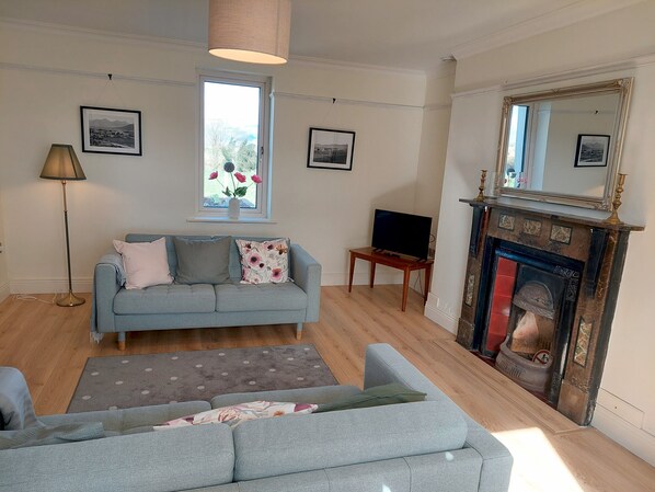 A living room with a sofa, a television on a stand, a fireplace, and a window with a view of trees.