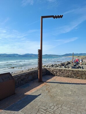 Un paseo marítimo con una escultura metálica, un muro de piedra y una playa al fondo.
