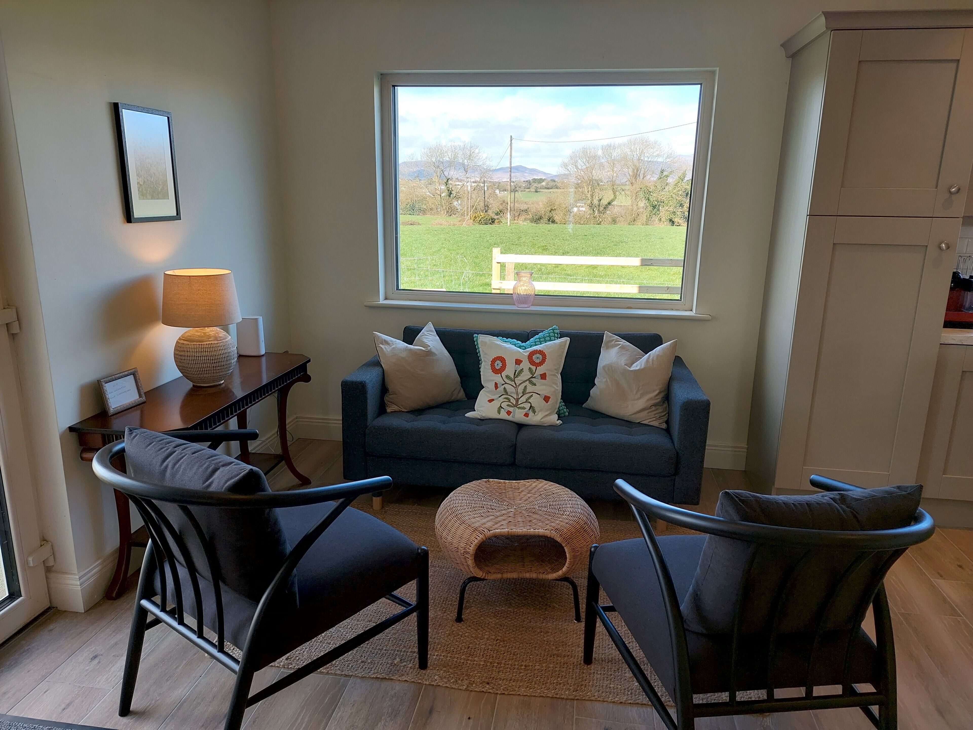 A cozy seating area with a sofa, two armchairs, a small table, and a lamp. A window offers a view of a green landscape.