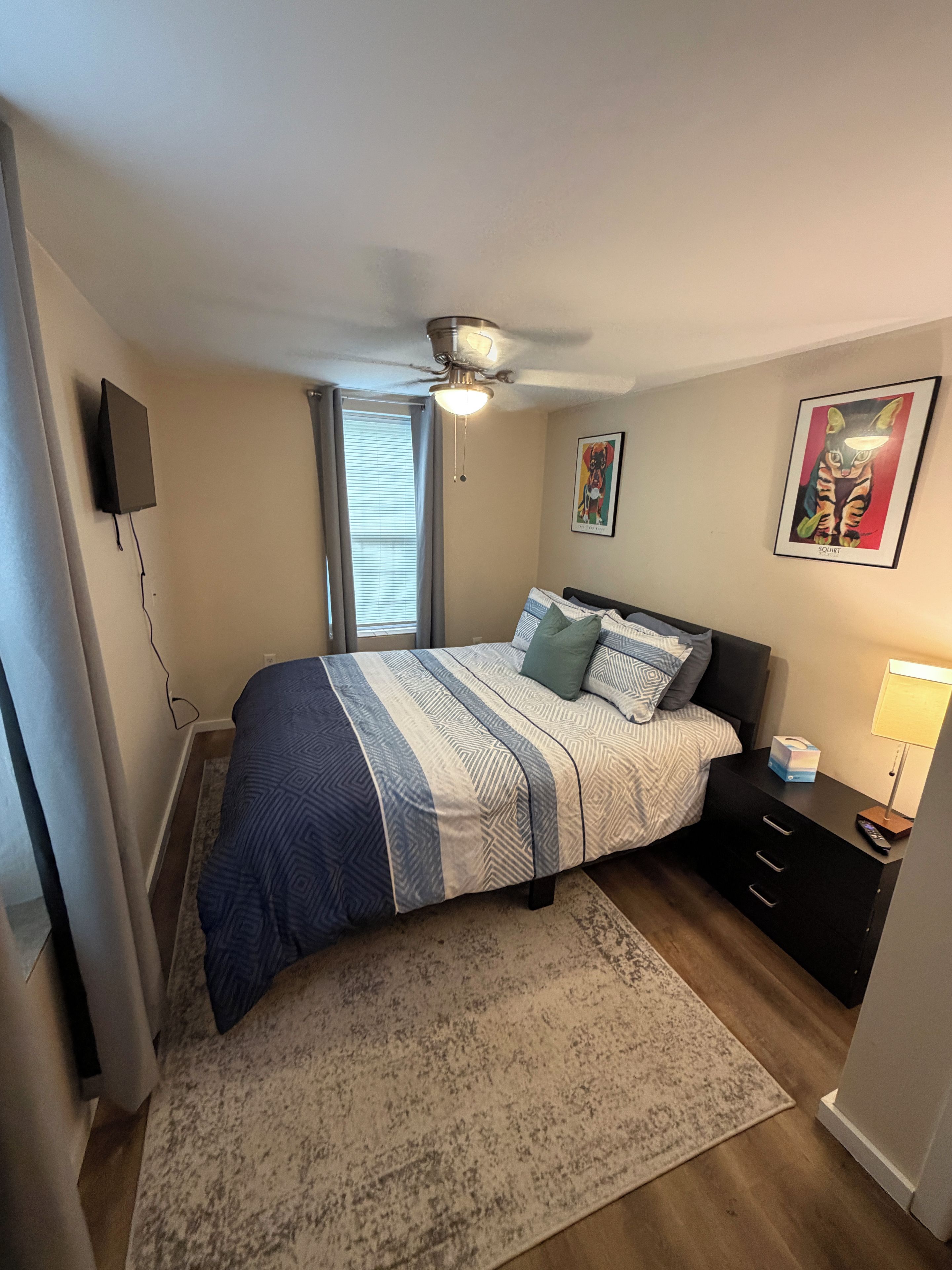 A bedroom with a bed, a nightstand, a lamp, a ceiling fan, and a window with blinds.