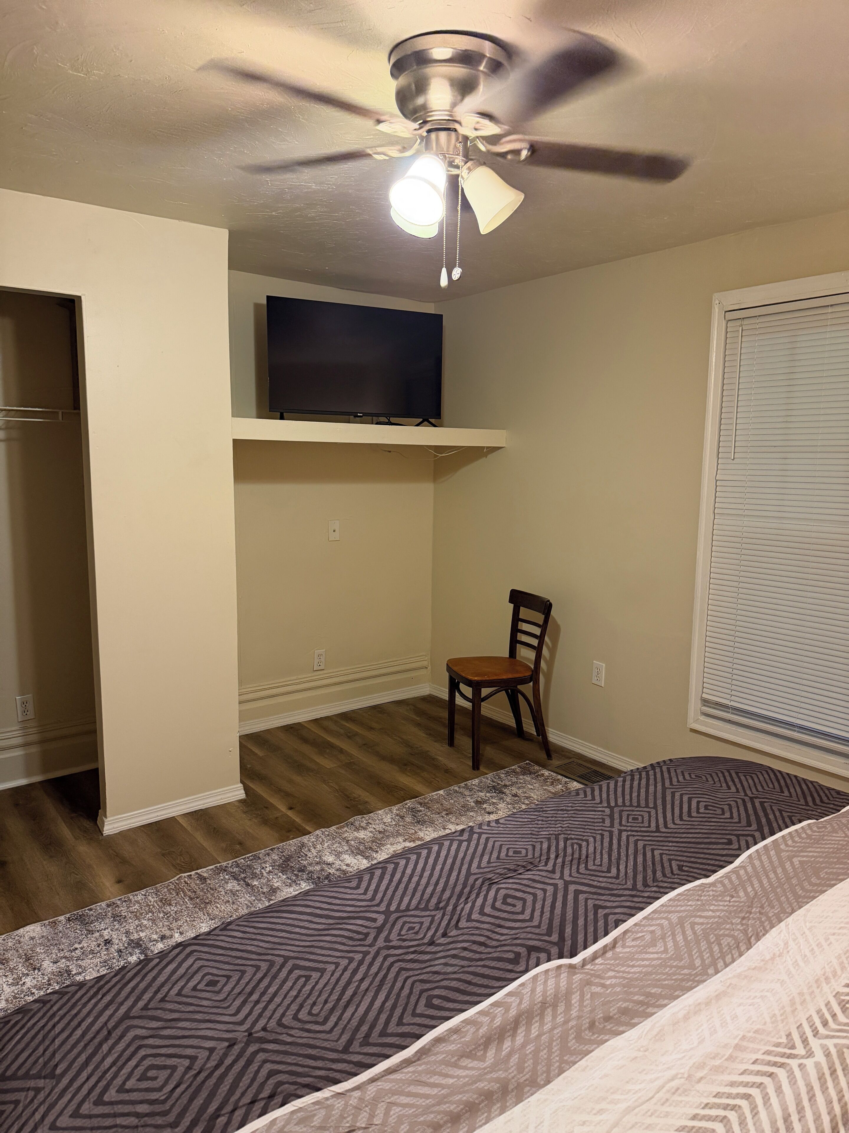 A bedroom with a ceiling fan, a flat-screen TV on a shelf, a chair, and a patterned rug.