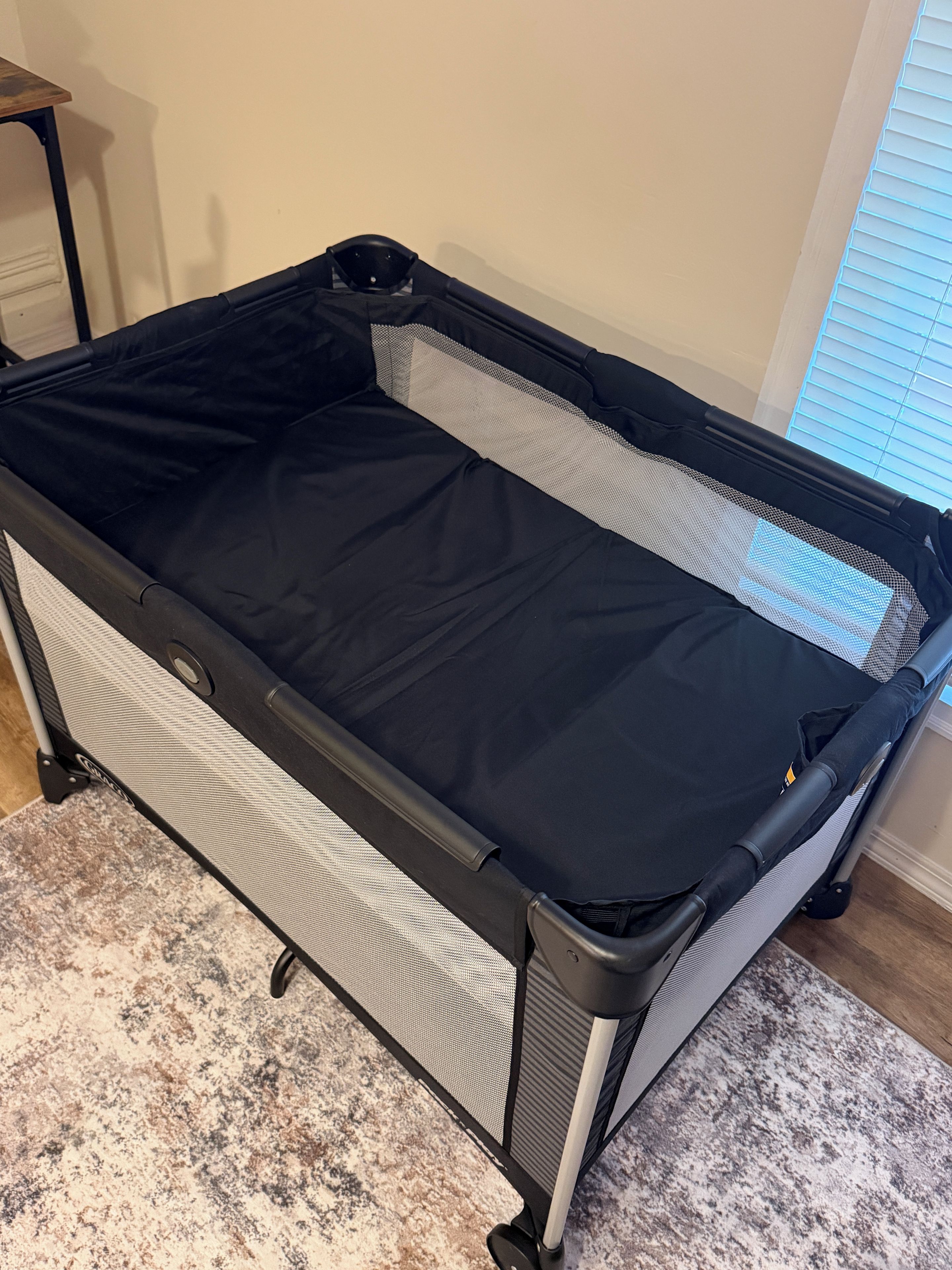 A portable crib with a mesh side and a solid black mattress, placed on a patterned rug.