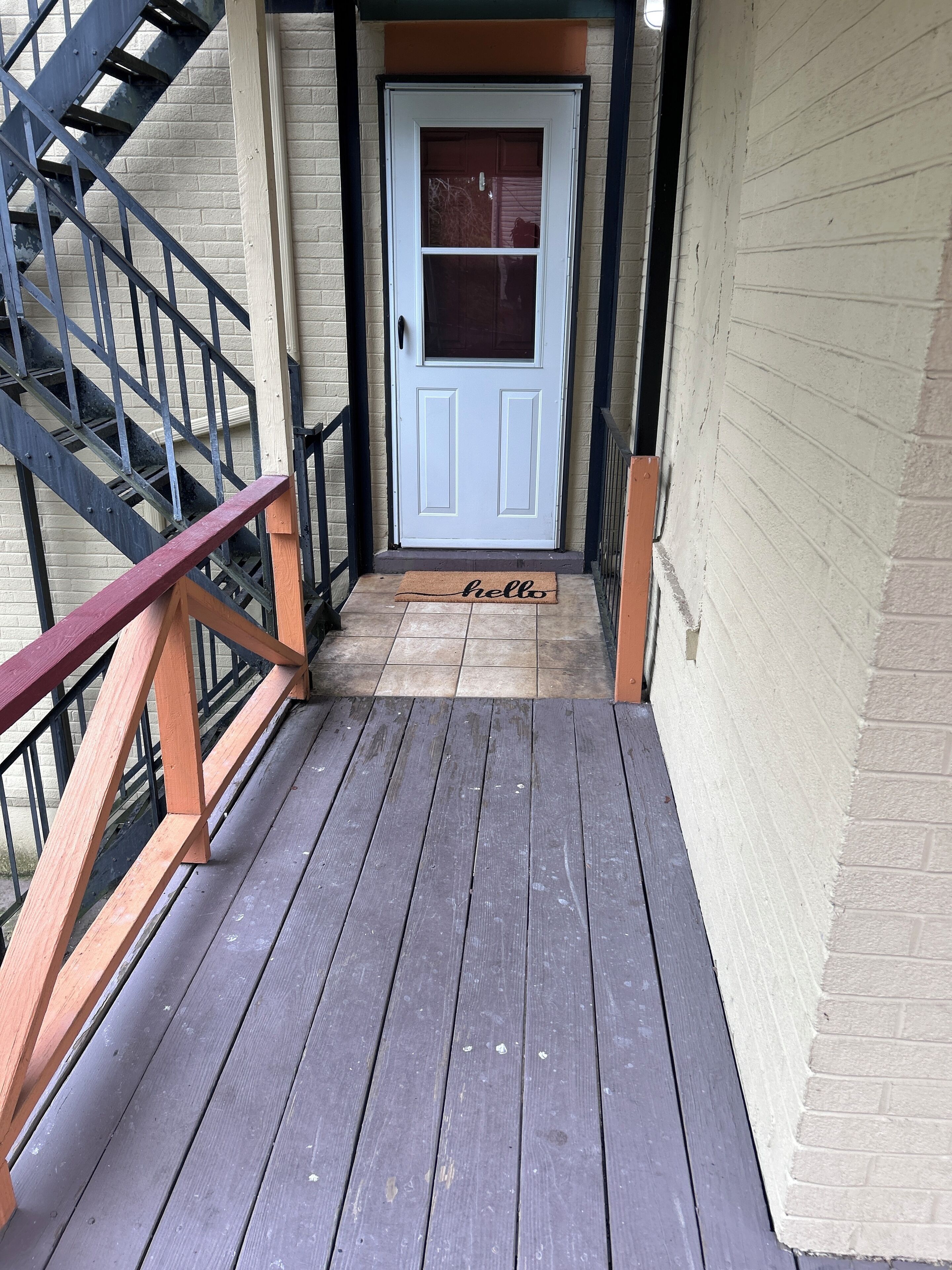 A wooden deck leading to a white door with a window, a metal staircase on the left, and a doormat with the word 'hello'.