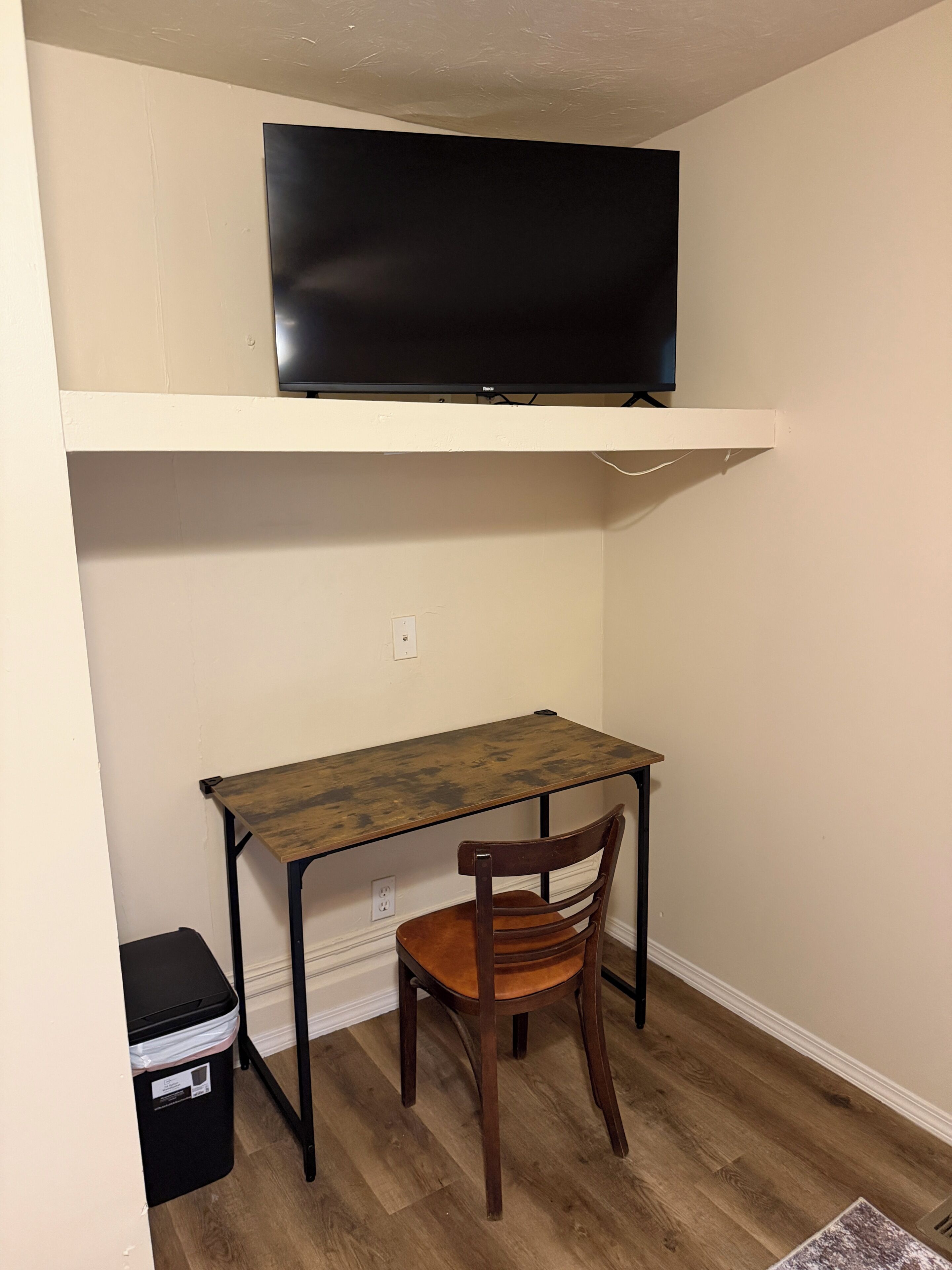 A small room with a wooden desk, a chair, a black trash bin, and a mounted television on the wall.