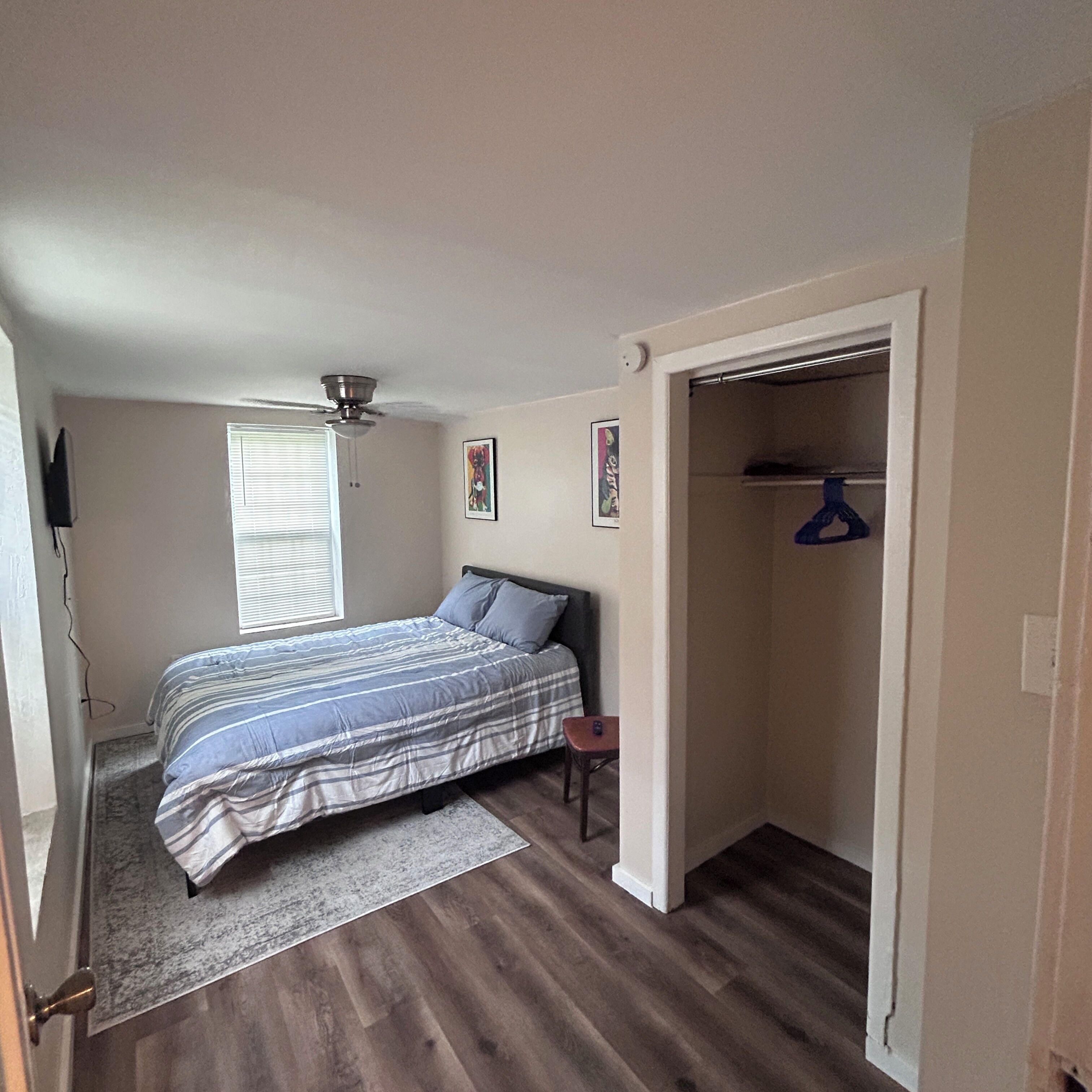 A bedroom with a bed, a ceiling fan, a window with blinds, a door leading to a closet, and a small table.