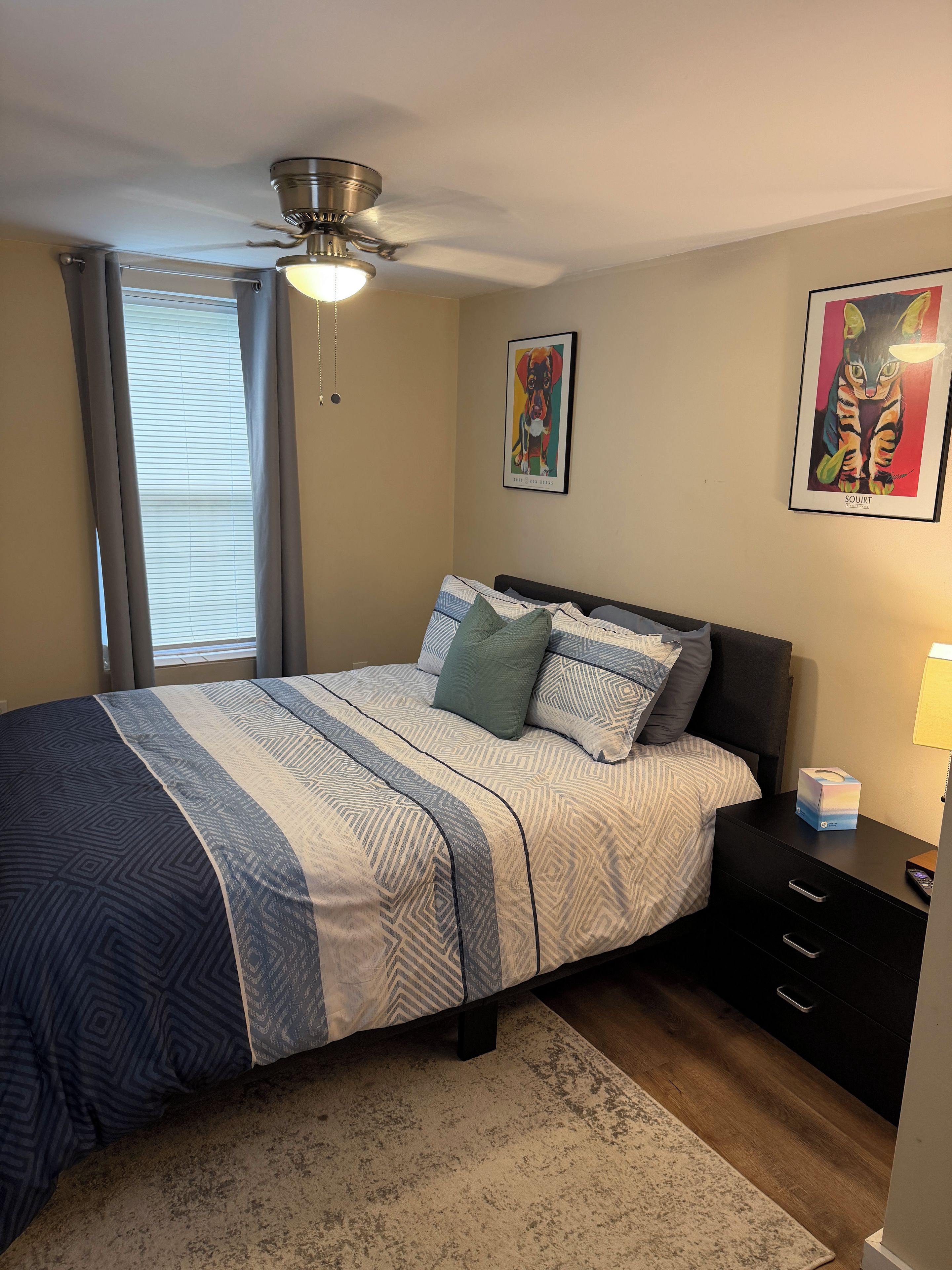 A bedroom with a bed, a nightstand, a drawer unit, a ceiling fan, and two wall art pieces.