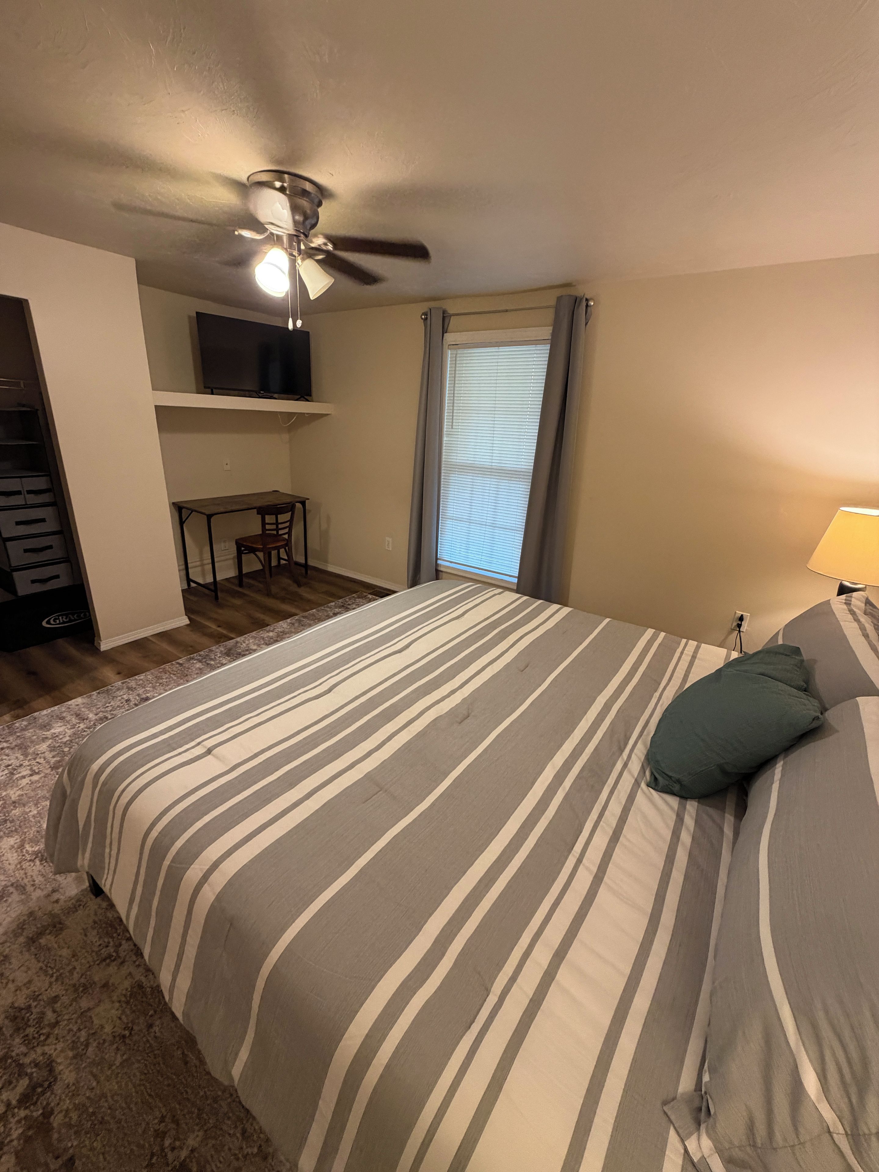 A bedroom with a bed, a desk, a chair, a ceiling fan, and a window with curtains.