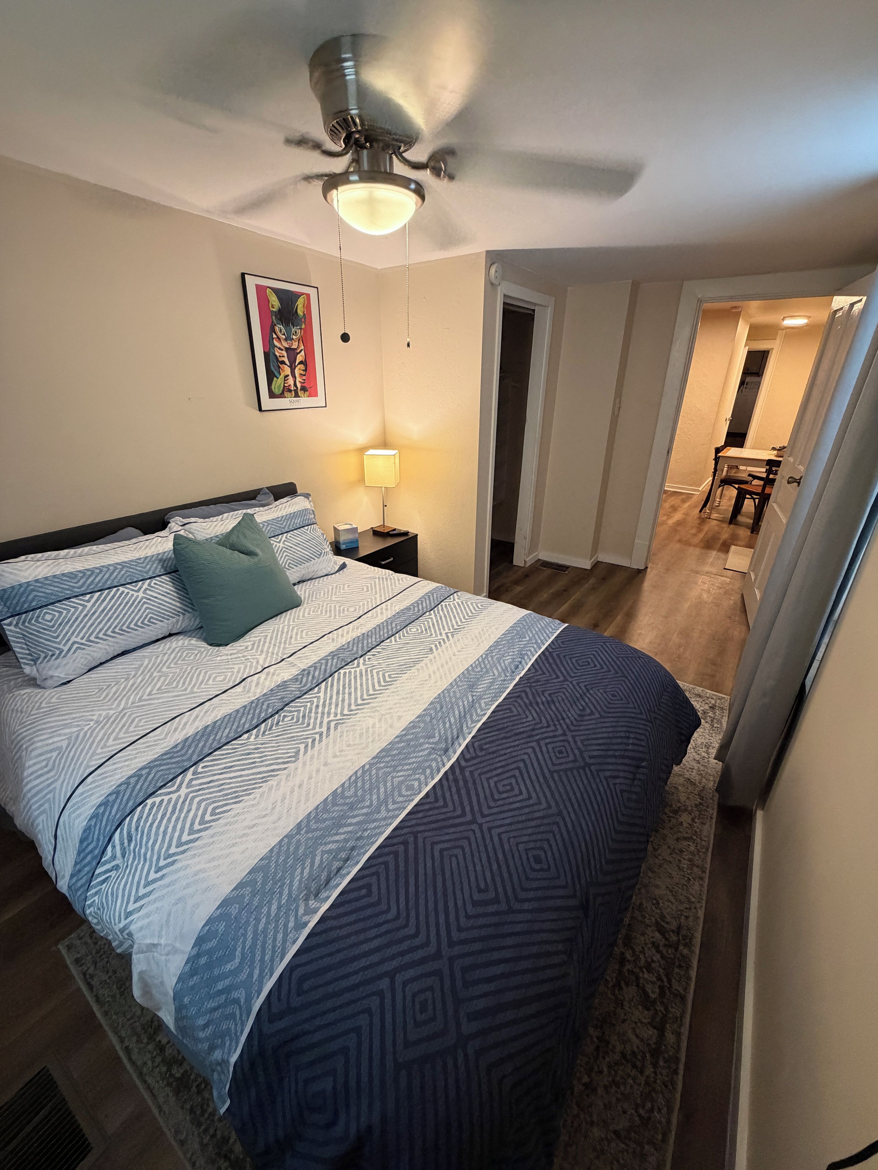 A bedroom with a bed, a nightstand, a lamp, a ceiling fan, and a door leading to another room.