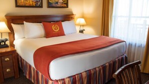 Ein ordentlich bezogenes Bett mit roter Tagesdecke, einem Holz-Kopfteil und einem gestreiften Bettschlitten. Auf dem Nachttisch stehen zwei Nachttischlampen und eine kleine Statue.