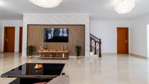 A spacious lobby with a flat-screen TV mounted on a wall, a wooden console table, two potted plants, a staircase with ornate railings, and a chandelier. - Spacious 5-Bedroom Villa with Garden Near Lisbon ,Sesimbra& beautiful beaches (Quinta Do Conde)