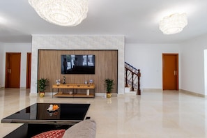 A spacious lobby with a flat-screen TV mounted on a wall, a wooden console table, two potted plants, a staircase with ornate railings, and a chandelier. - Spacious 5-Bedroom Villa with Garden Near Lisbon ,Sesimbra& beautiful beaches (Quinta Do Conde)