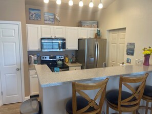 A kitchen with white cabinets, a stainless steel refrigerator, a microwave, and a stove. There are two wooden chairs with black cushions.