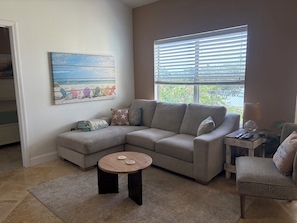 A living room with a sectional sofa, a small round table, and a painting of beach chairs. - Two Bed Two Bath Seminole Stay (Seminole)