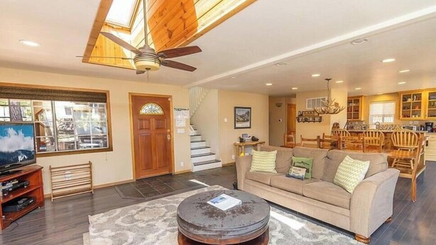 A spacious living room with a large round coffee table, a sofa, and a television. There is a staircase leading to an upper level, and a kitchen area is visible in the background. - Lakefront 4BR, Dock, Hot Tub, EV Charger, Dogs OK (Big Bear Lake)