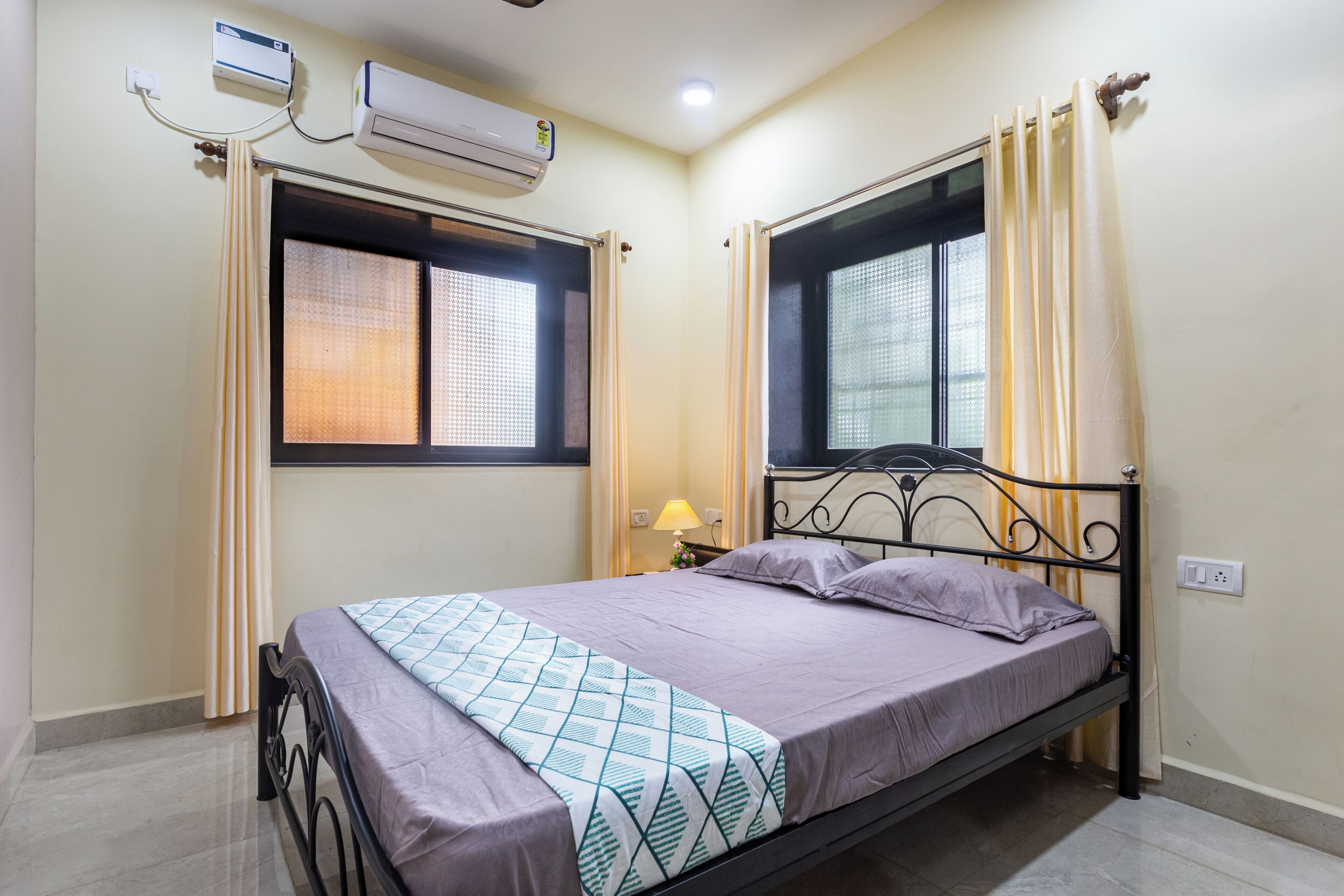 A single bed with a metal frame, a window with a view, and an air conditioning unit on the wall.