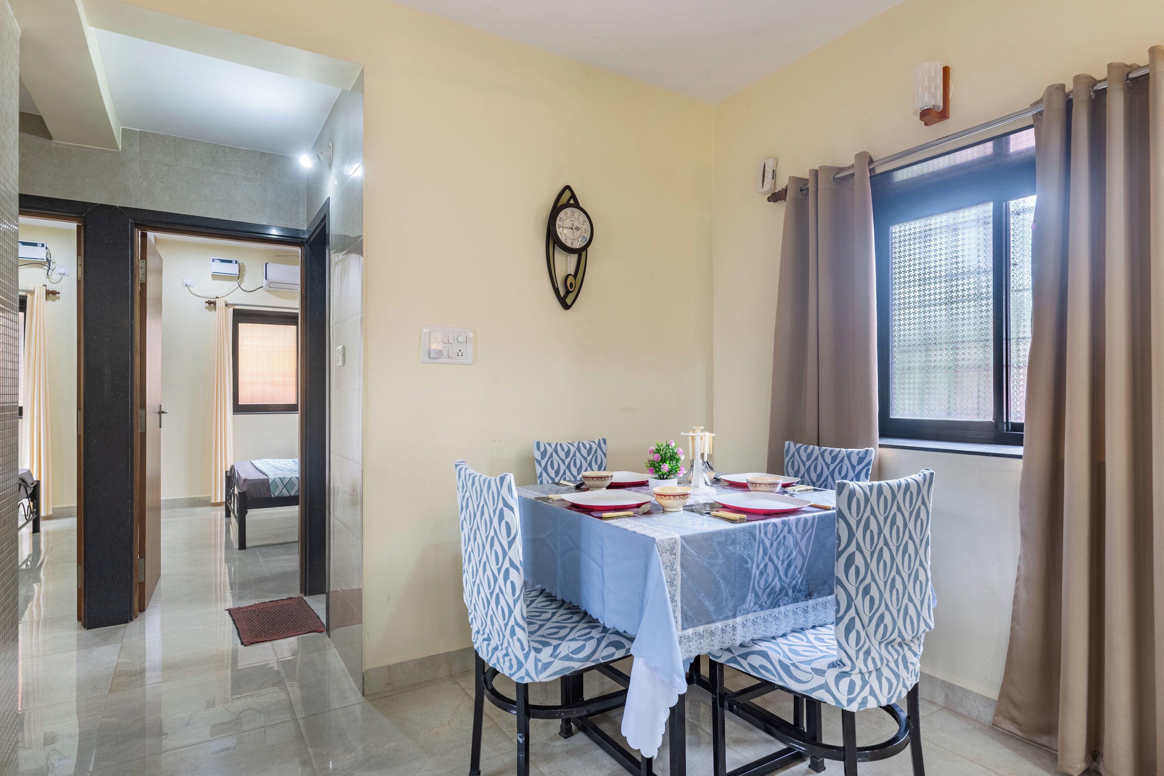 A dining area with a table set for four, patterned chairs, a wall clock, and a window with curtains.