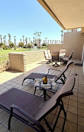 Eine Terrasse im Freien mit Lounge-Möbeln und einem Tisch, eine Ziegelwand und Blick auf eine Rasenfläche mit Palmen.