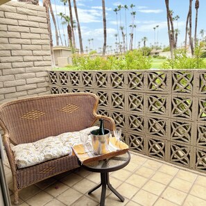 Un canapé en osier agrémenté d’un coussin à motifs, une petite table ronde avec un verre et une bouteille, le tout placé sur une terrasse carrelée, avec une clôture décorative et des palmiers en arrière-plan.