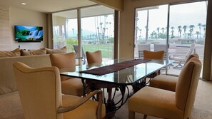 A modern hotel room with a glass dining table, beige upholstered chairs, a flat-screen TV mounted on the wall, and a view of palm trees through large windows. - Indian Wells 3 bed retreat with incredible views, multiple pools and tennis (Indian Wells)