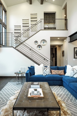 Una sala de estar moderna con un sofá seccional azul, una mesa de centro con libros, una escalera con barandilla y decoración en la pared.