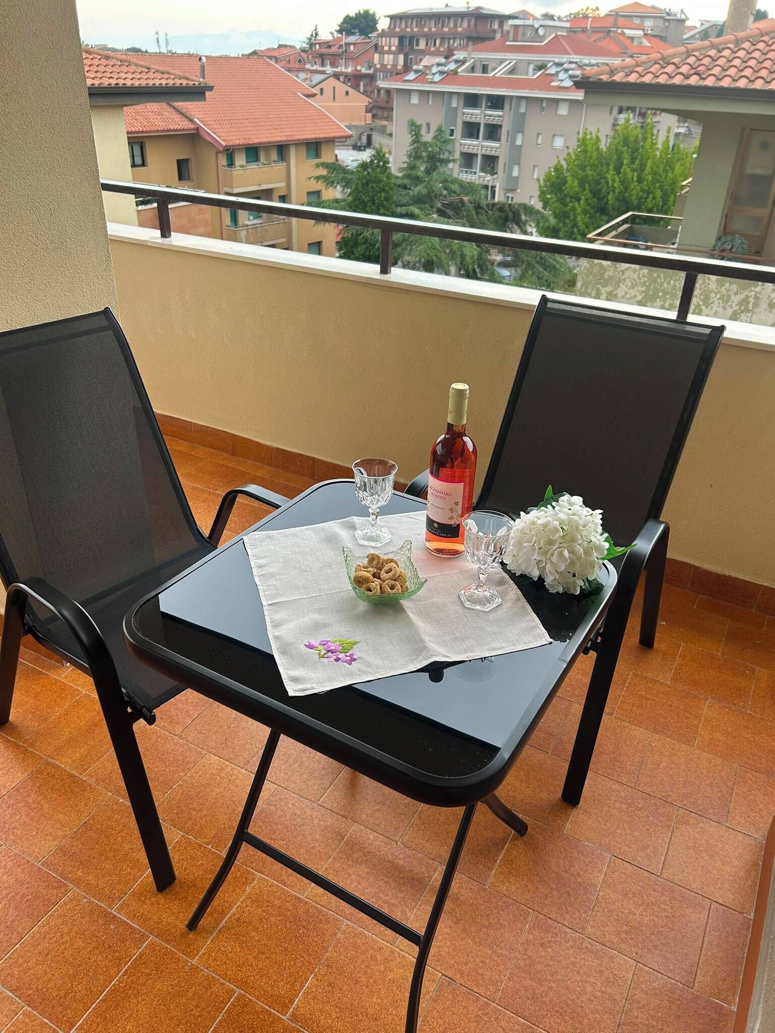 Un balcone con un tavolo apparecchiato per due, con una bottiglia di vino, un bicchiere, una ciotola di stuzzichini e un vaso di fiori.