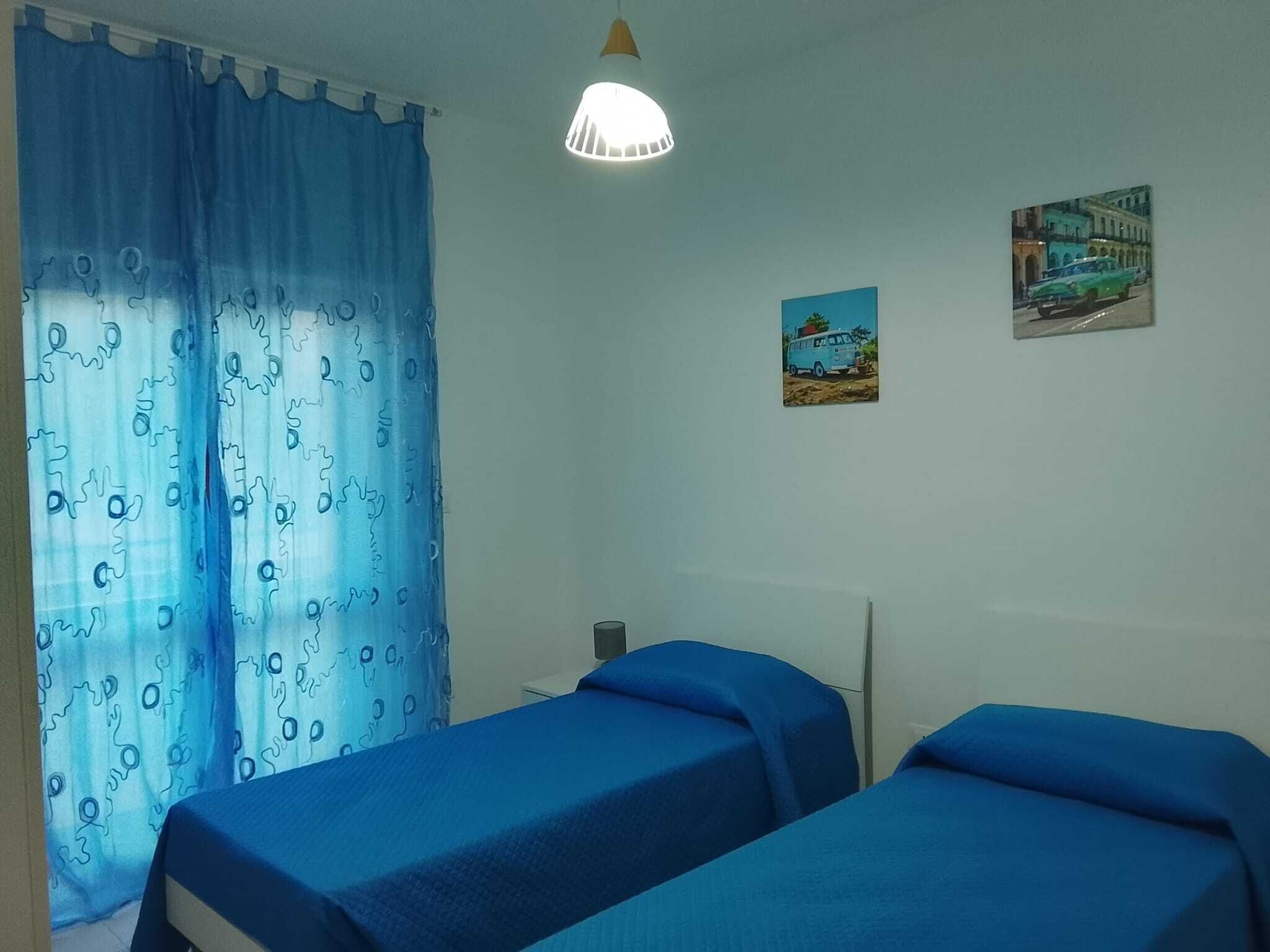 Una stanza con due letti, una finestra con tende azzurre, una lampada a parete e due piccoli quadri.