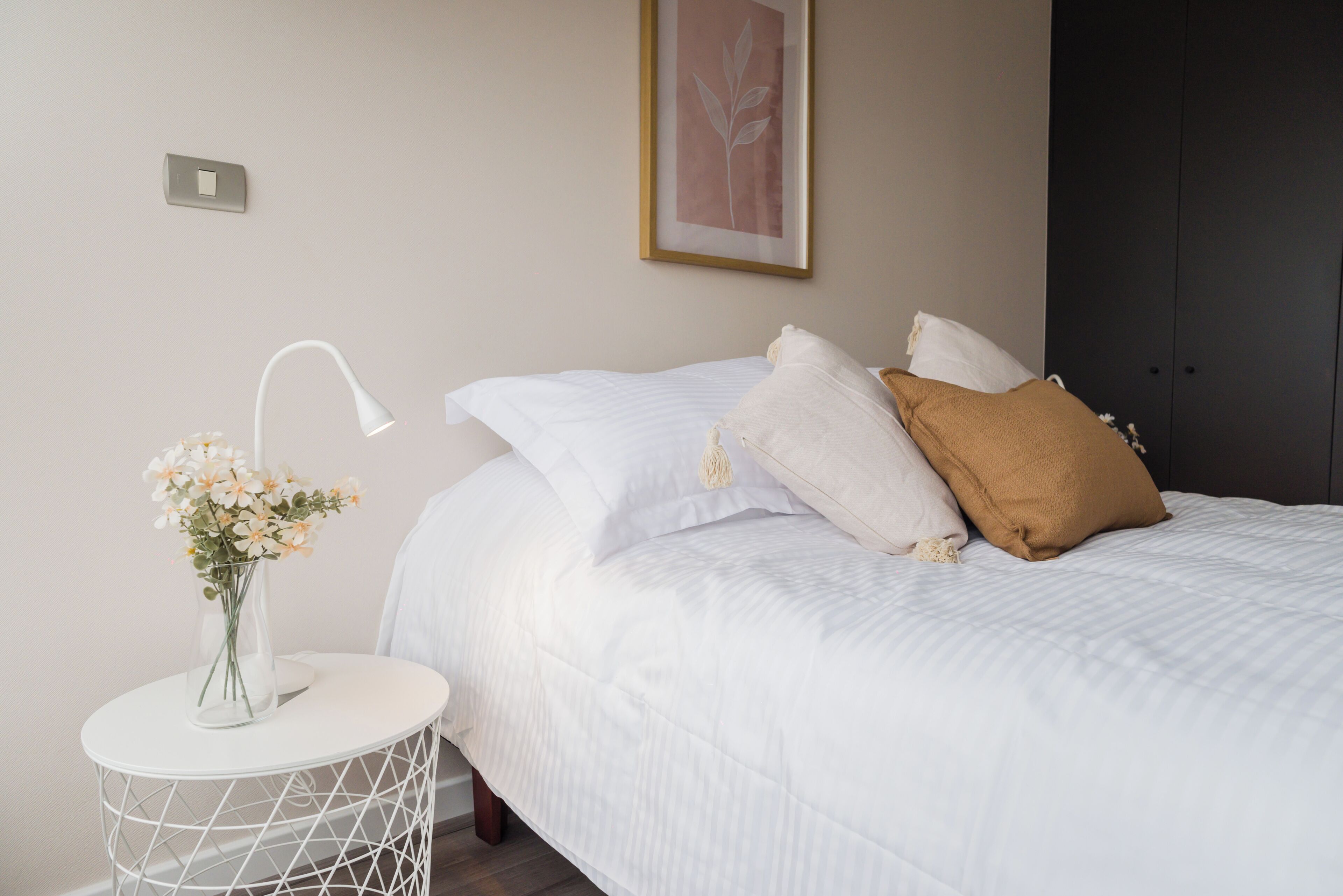A neatly made bed with white bedding and a brown pillow, a white bedside table with a lamp and a vase of flowers, and a framed picture on the wall.