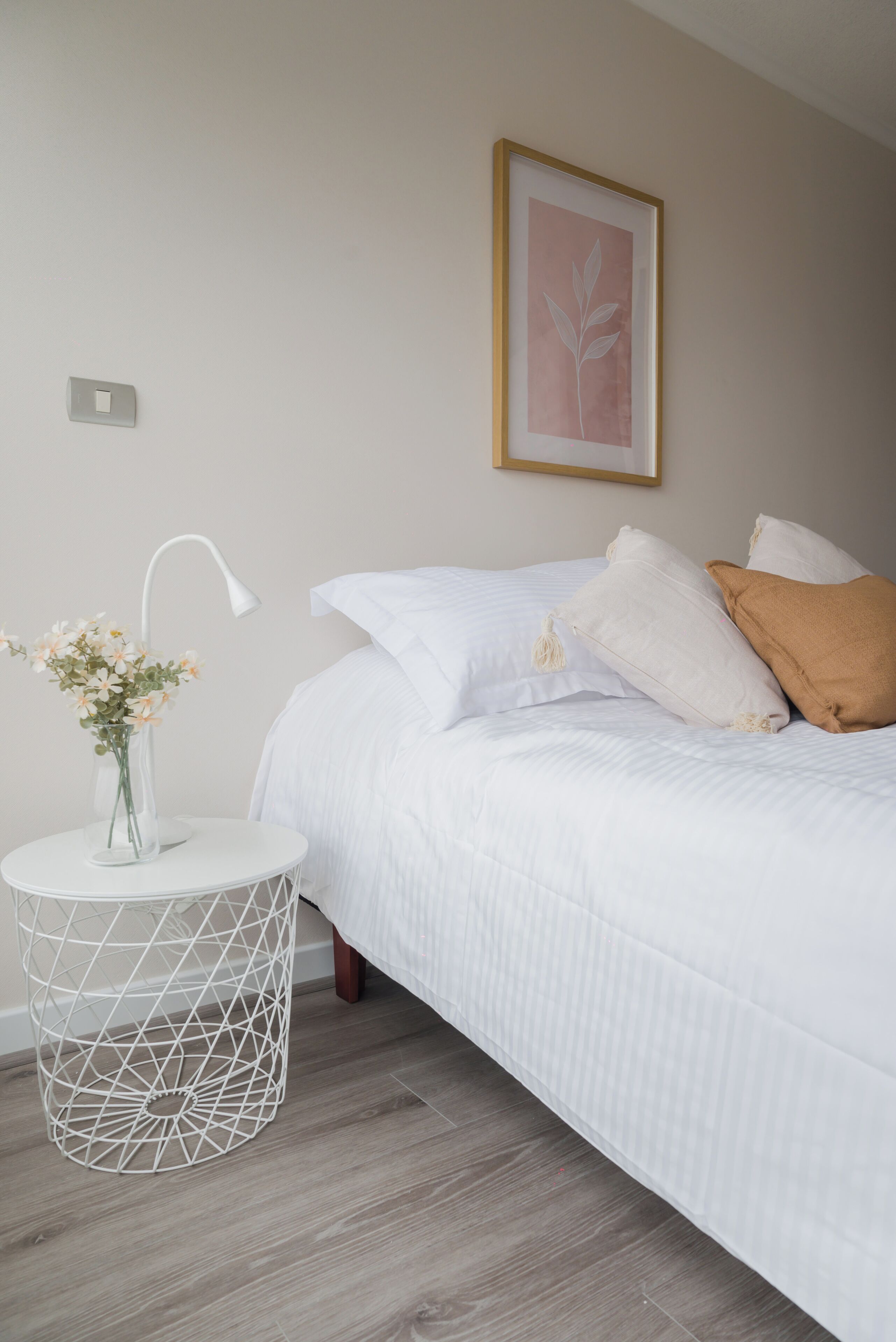 A bedroom with a bed, a bedside table with a vase of flowers, a wall-mounted light switch, and a framed picture on the wall.