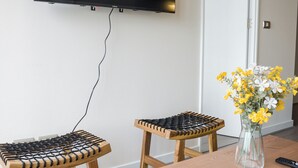 A room with a flat-screen TV mounted on the wall, two wooden stools with black rope seating, a wooden table, and a vase with yellow and white flowers. - Seminario Apartments by Andes STR (Ñuñoa)