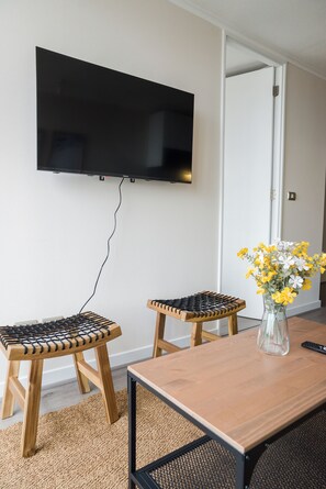 A room with a flat-screen TV mounted on the wall, two wooden stools with black rope seating, a wooden table, and a vase with yellow and white flowers. - Seminario Apartments by Andes STR (Ñuñoa)