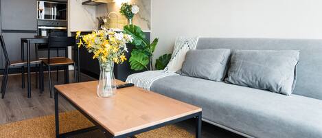 Una sala moderna con un sofá gris, una mesa de centro de madera con un jarrón de flores amarillas, una cocina con armarios blancos y una mesa de comedor con sillas.