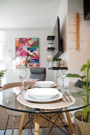 Een eetruimte met een glazen tafel, borden, bestek en een kleurrijk abstract schilderij aan de muur.