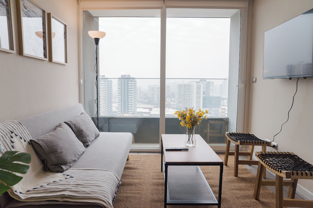 A modern living room with a sofa, a coffee table, a small stool, a TV mounted on the wall, and a view of the cityscape through large windows.