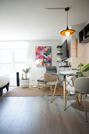 A modern living room with a dining area, a colorful abstract painting, and a large window. - Seminario Apartments by Andes STR (Ñuñoa)