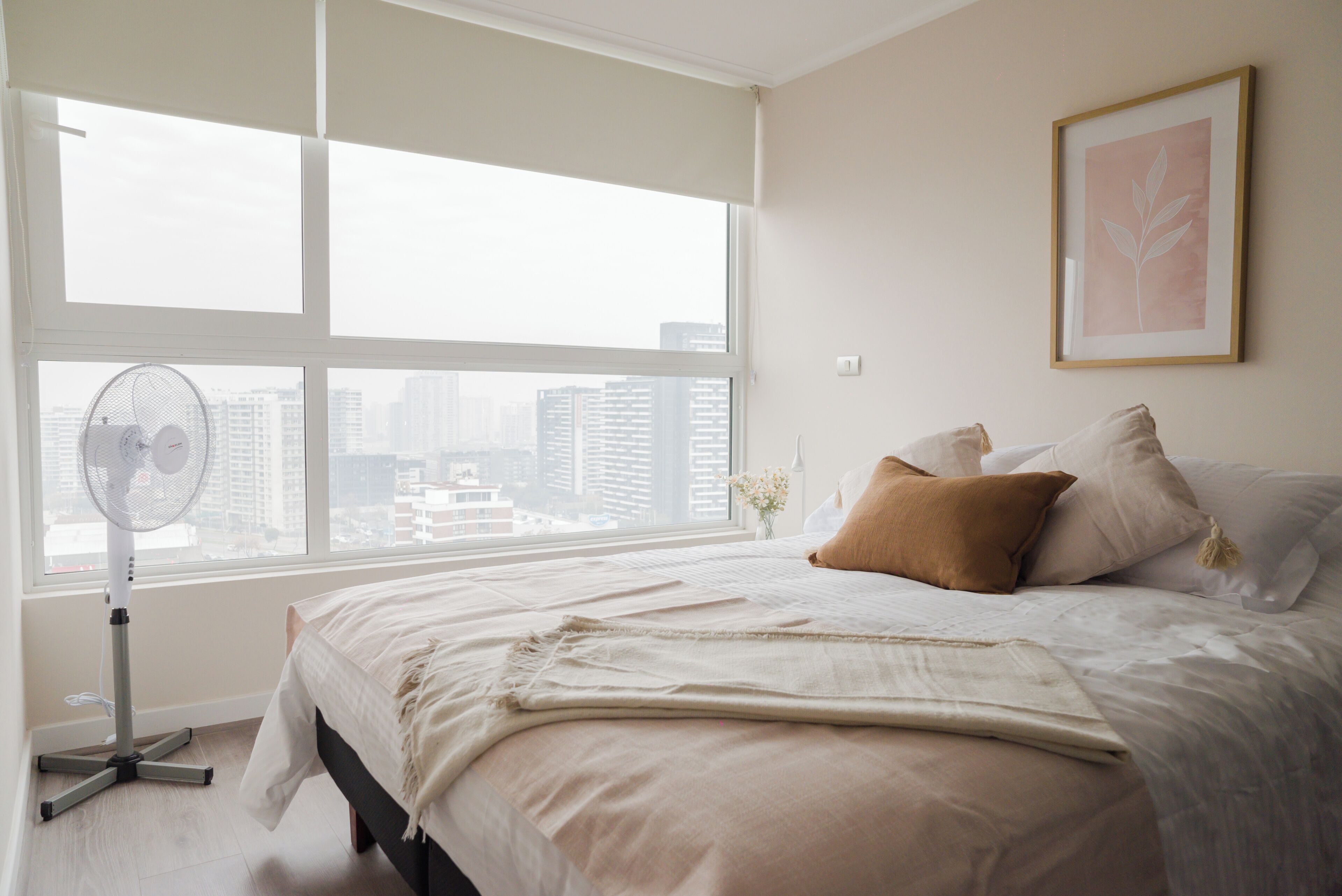 Un dormitorio con un ventanal grande, un ventilador de pie y una cama con almohadas.