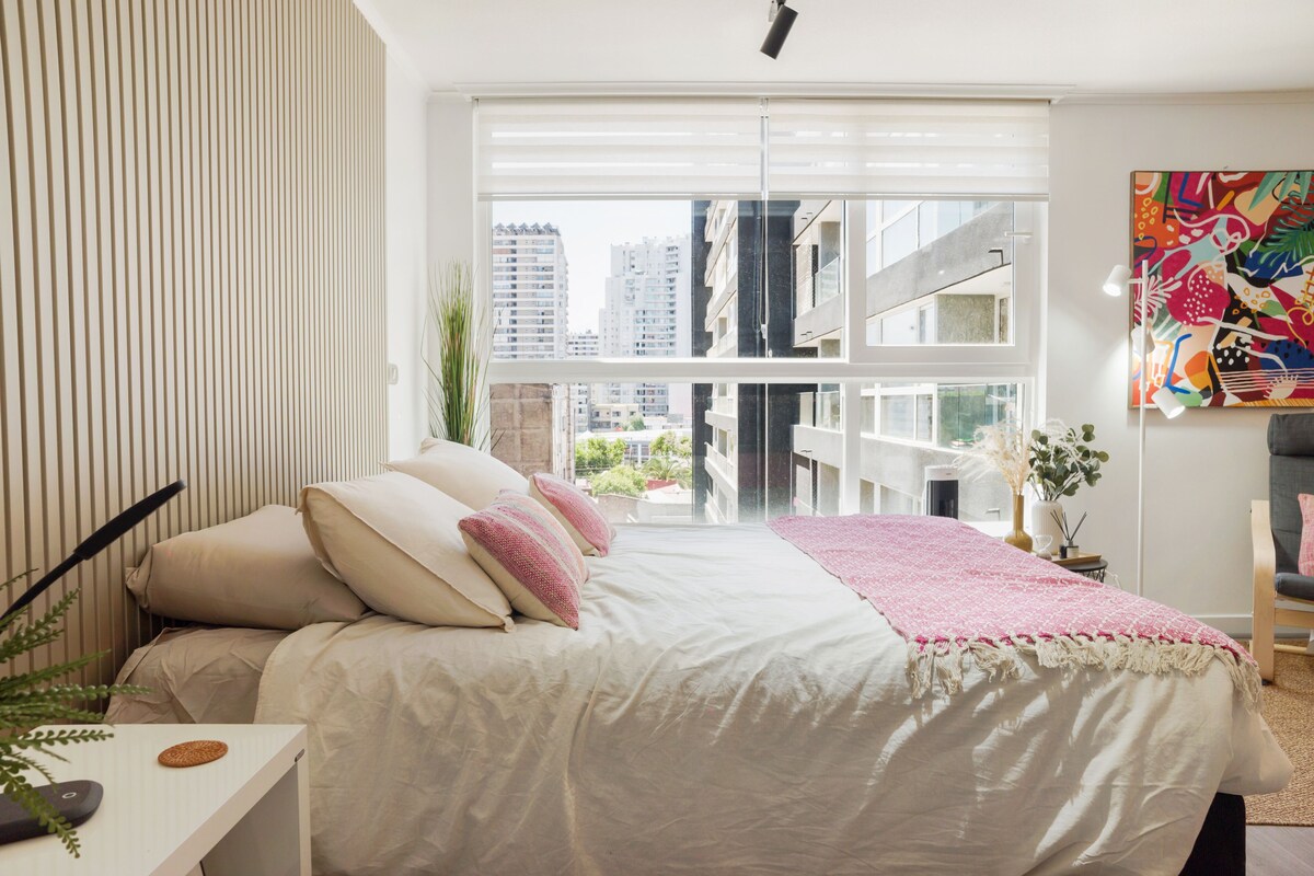 A modern bedroom with a large bed, a view of the city, and a colorful abstract painting on the wall.