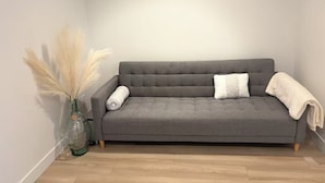 Un sofá gris con dos almohadas y una manta, un jarrón de vidrio con plumas de pampa, y un piso de madera.