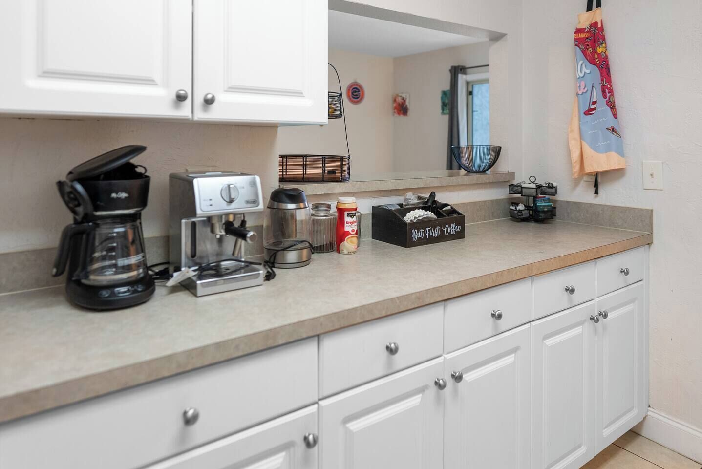 A kitchen with white cabinets, a coffee maker, a kettle, a coffee grinder, and a coffee bean container.