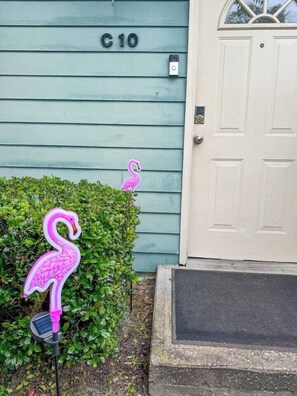 Une maison avec le numéro « C10 » sur le mur, une porte beige et un petit jardin agrémenté d’une décoration en forme de flamant rose.