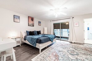 A bedroom with a bed, bedside tables, a desk, a ceiling fan, and a view of the outside balcony. - Spacious 4BR | Near UF & Shands | Pool + Gym (Gainesville)
